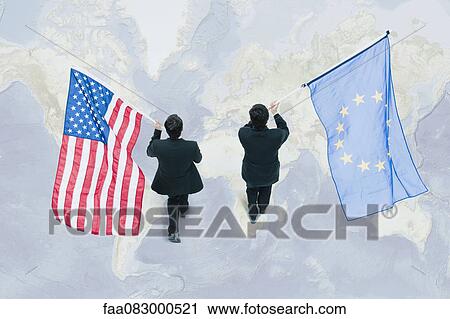 Stock Image - Businessman walking on top of world map, carrying American and European Union flags. Fotosearch
