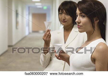 Stock Photo - Businesswomen taking break and drink from disposable cups in hallway. Fotosearch