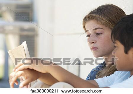 Stock Image - Children sitting together outdoors, looking at book. Fotosearch