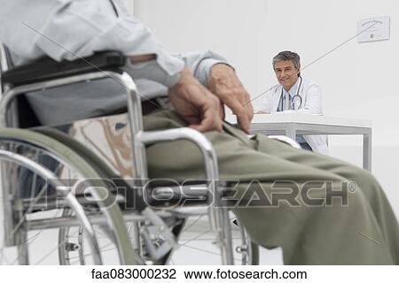 Stock Image - Doctor working in office, patient in wheelchair in foreground. Fotosearch