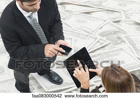Stock Image - Executives exchanging information on wireless devices, pile of money in background. Fotosearch