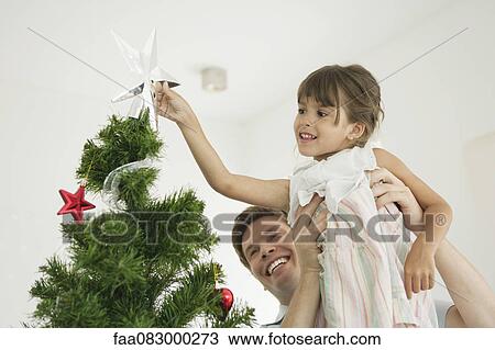 Stock Image - Father carrying little girl putting star on tip of Christmas tree. Fotosearch
