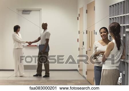 Stock Image - Female office workers chatting by filing shelves, people shaking hands in background. Fotosearch