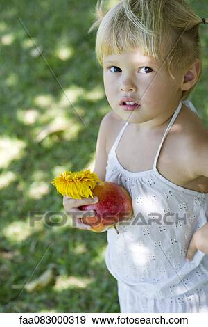 Stock Photo - Little girl standing outdoors, holding apple and flower. Fotosearch