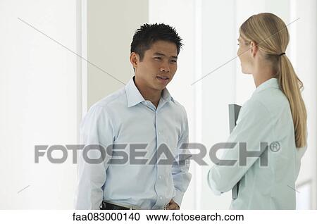 Stock Image - Mid-adult man talking to female colleague in office corridor. Fotosearch