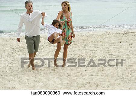 Stock Image - Parents swinging young daughter as they walk on beach. Fotosearch