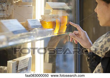 Stock Image - Woman looking at jars of honey in shop window. Fotosearch