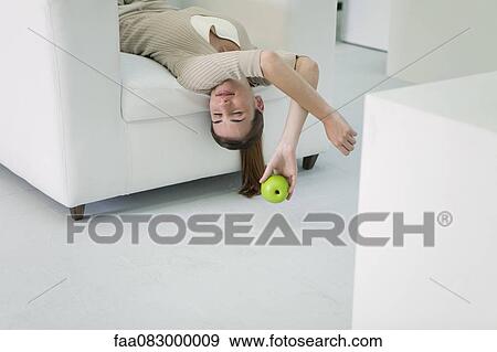 Stock Photo - Woman lying upside down on sofa, holding apple. Fotosearch