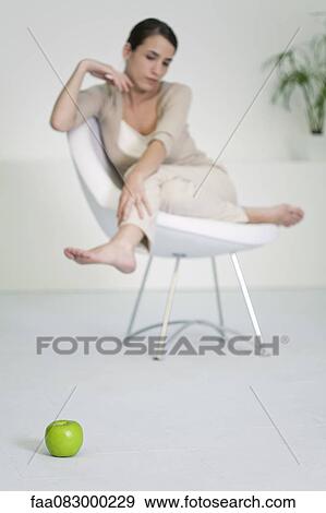 Stock Photo - Woman relaxing in chair, looking at apple on floor in foreground. Fotosearch