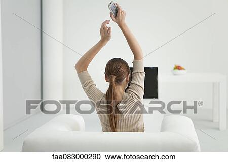 Stock Image - Woman watching TV with arms raised in air, rear view. Fotosearch