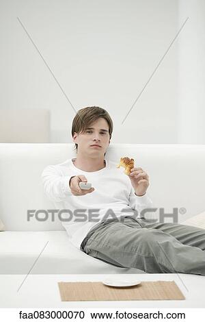 Stock Image - Young man eating croissant while watching TV. Fotosearch