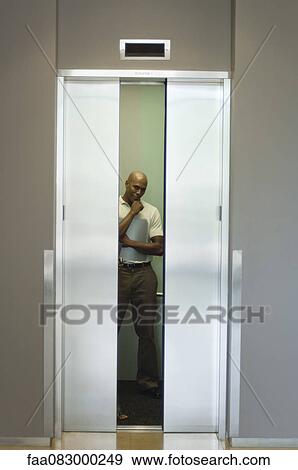 Stock Photo - Young man standing in elevator with hand under chin, doors closing. Fotosearch