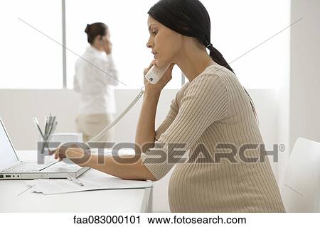 Stock Image - Young pregnant woman talking on landline phone in office, backlit. Fotosearch