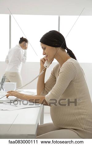 Stock Image - Young pregnant woman talking on landline phone in office, backlit. Fotosearch