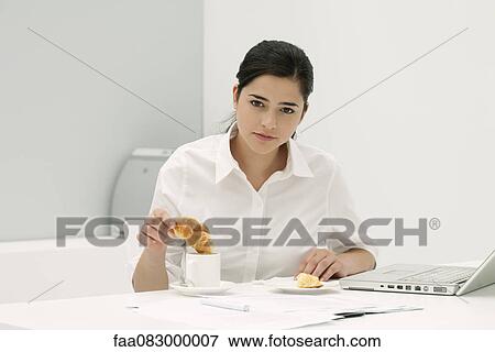 Stock Photo - Young woman dipping croissant in coffee at desk in office. Fotosearch