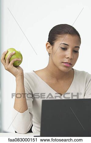 Stock Image - Young woman eating apple at desk. Fotosearch
