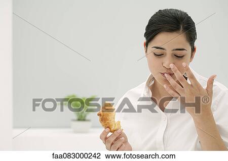 Stock Image - Young woman licking finger while eating croissant. Fotosearch