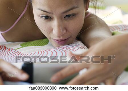 Stock Image - Young woman lying down, looking at cell phone. Fotosearch