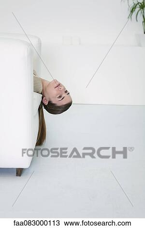 Stock Image - Young woman lying on sofa with head dangling, daydreaming. Fotosearch