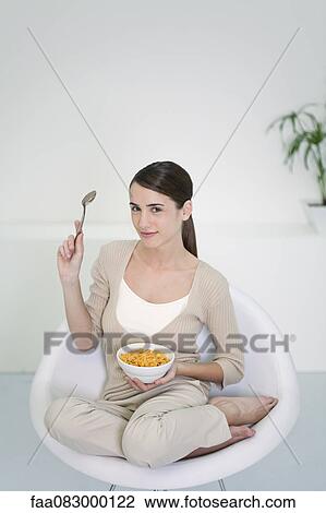 Stock Image - Young woman sitting in chair with bowl of cereal. Fotosearch