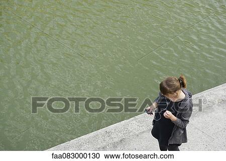 Stock Image - Young woman walking along canal, listening to MP3 player, elevated view. Fotosearch