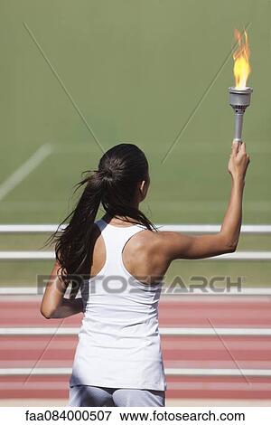 Stock Photo - Female athlete holding up flaming torch, rear view. Fotosearch