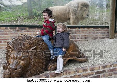 Stock Photograph - Children playing on lion statue in front of lion enclosure at the zoo. Fotosearch