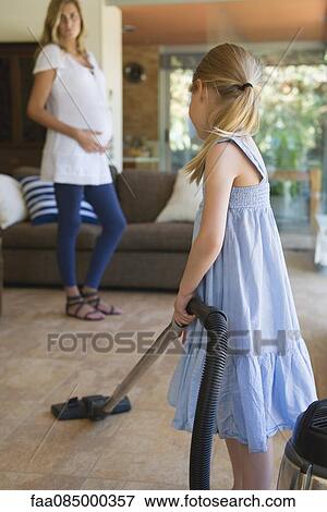 Stock Photo - Little girl vacuuming, pregnant mother in background. Fotosearch