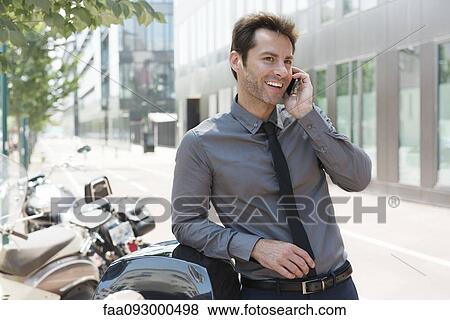 Businessman talking on cell phone in city, leaning against motor scooters View Large Photo Image Stock Photo - Businessman talking on cell phone in city, leaning against motor scooters. Fotosearch