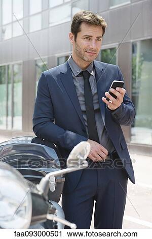 Stock Image - Businessman using smartphone in city, leaning against motor scooters. Fotosearch