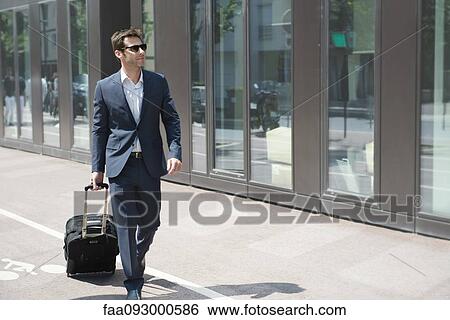 Stock Photograph - Businessman with sunglasses pulling luggage, walking in city. Fotosearch
