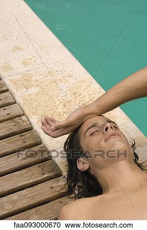 Stock Image - Man lying on back next to pool, touching forehead with back of hand. Fotosearch