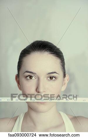 Picture - Woman holding measuring tape across mouth, close-up. Fotosearch