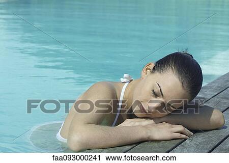 Stock Image - Woman in pool leaning against deck, resting head on arms. Fotosearch