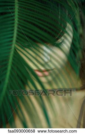 Stock Photo - Woman standing behind leaves, eyes closed, selective focus. Fotosearch