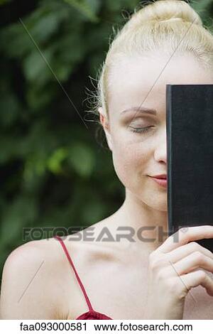 Stock Image - Young woman holding book in front of face, eyes closed. Fotosearch