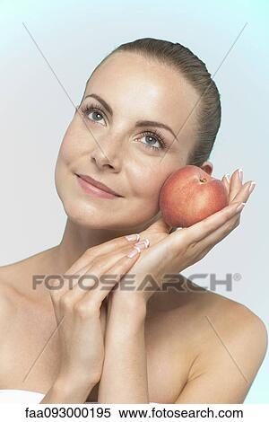 Stock Photography - Young woman holding peach to cheek. Fotosearch
