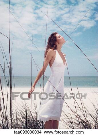 Picture - Young woman in dress standing in dune grass with head back, facing wind. Fotosearch