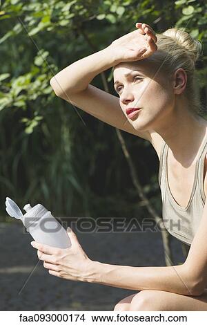 Young woman sitting outdoors with water bottle, wiping forehead with back of hand View Large Photo Image Picture - Young woman sitting outdoors with water bottle, wiping forehead with back of hand. Fotosearch
