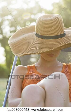 Stock Image - Young woman sunbathing outdoors, face covered by sun hat. Fotosearch