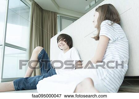 Stock Photo - Brother and sister sitting on floor, leaning against back of sofa. Fotosearch