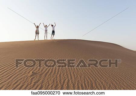 Stock Image - Children jumping on top of dune with arms raised in air. Fotosearch