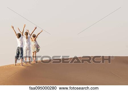Stock Image - Children standing in desert with arms raised in air. Fotosearch