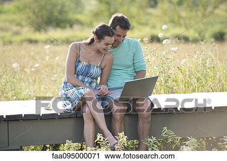 Stock Image - Couple sitting on countryside boardwalk with laptop computer. Fotosearch