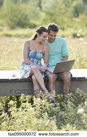 Stock Image - Couple sitting on countryside boardwalk using laptop computer. Fotosearch