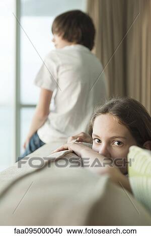 Girl sitting on sofa with head resting on arm, brother looking out window in background View Large Photo Image Stock Image - Girl sitting on sofa with head resting on arm, brother looking out window in background. Fotosearch