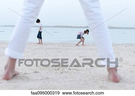 Stock Photo - Girl standing on beach, watching siblings at water's edge. Fotosearch
