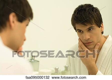 Stock Image - Man in bathrobe looking in mirror, touching face. Fotosearch