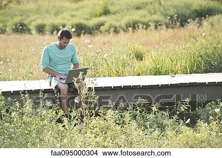 Picture - Man using laptop computer while sitting out in countryside. Fotosearch