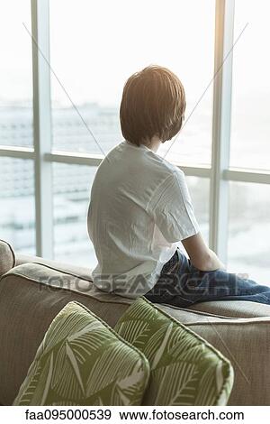 Stock Photo - Teenage boy sitting on edge of sofa, looking out window, rear view. Fotosearch